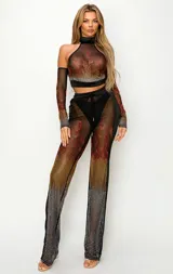 Conjunto de dos piezas compuesto por un top de cuello alto con mangas largas integradas y un pantalón de tiro alto. Ambas prendas están confeccionadas en tejido de malla transparente con un diseño estampado de llamas en tonos rojos, naranjas y dorados, decorado con pedrería brillante que se intensifica hacia el ruedo.
