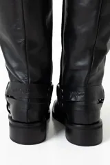 Botas de caña alta confeccionadas en cuero negro, con diseño de estilo western que incluye una correa decorativa con argollas metálicas y remaches en el tobillo. Presentan una puntera cuadrada y un taco bajo cuadrado.