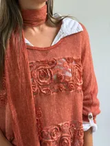Sweater de punto color naranja con diseño holgado, que incorpora franjas horizontales de encaje con motivos florales. Presenta mangas tres cuartos con puños abotonados y se complementa con una bufanda del mismo tejido y color.