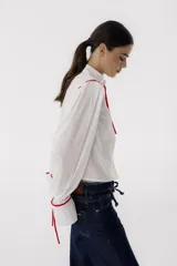 Camisa blanca de corte cropped con cuello mao y mangas largas abullonadas. Presenta detalles de ribetes rojos en cuello, mangas y hombros, y lazos rojos en los puños y en el cuello.