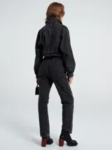 Mono de denim negro con cierre frontal de botones, cintura elástica y detalle de flecos en la parte superior delantera. Presenta mangas largas con puños elásticos y corte recto en las piernas.