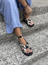 Sandalia de cuero color plateado con plataforma. Presenta tiras cruzadas en la parte delantera, una tira con agarre para el dedo gordo y una pulsera ajustable con hebilla en el tobillo.