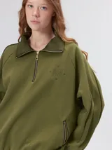 Buzo verde militar de algodón suave, de calce oversize, con cuello alto y cierre frontal parcial metalizado. Cuenta con bolsillos laterales con cierre y puños y ruedo elastizados.