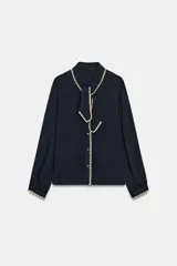 Blusa de manga larga en color azul marino, con cuello solapa y lazo frontal. Presenta detalles de pespuntes bordados en color crema a lo largo del cuello, lazo, cierre frontal y puños. Cierre frontal con botones.