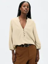 Blusa color beige claro, de manga larga con elástico en el puño, escote en V con cuello mao y tapeta corta con botones oscuros.