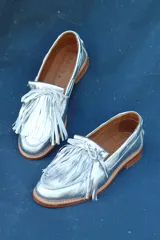 Mocasín plateado confeccionado en cuero metalizado, con flecos y detalles entrelazados.
