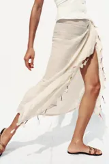 Pañuelo o foulard de tejido ligero color crudo o beige claro, con finas líneas verticales tejidas en un tono más claro. El borde inferior está decorado con borlas o pompones en tonos contrastantes como fucsia, morado y beige/dorado.