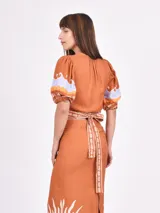 Blusa color ladrillo con escote cruzado, mangas cortas abullonadas con detalle de estampado de ondas en lila y naranja, y ribete de conchillas en escote, cintura y mangas.