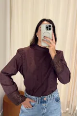 Blusa de manga larga color marrón oscuro, con cuello alto y diseño de encaje en los puños. Presenta un patrón de bordado inglés en toda la prenda, con mangas ligeramente abullonadas en los hombros.