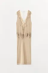 Vestido midi de color beige con escote en pico y tirantes anchos. Presenta un diseño con tejido plisado en la parte superior y aplicaciones de encaje a tono en el escote y la cintura. Cuenta con una tira ajustable con lazada en la espalda y forro interior.