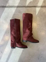 Bota de caña alta confeccionada en gamuza color bordó, con diseño de solapa lateral y cierre vertical. Presenta una silueta estructurada con taco bajo y suela plana.