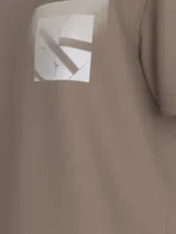 Remera de manga corta color topo (marrón grisáceo) con cuello redondo y bolsillo en el pecho. El bolsillo presenta un estampado difuminado del monograma de Calvin Klein Jeans.