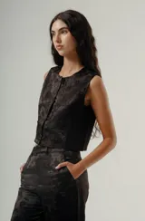 Top negro sin mangas, estilo chaleco corto, con cuello redondo y cierre frontal de botones. Presenta un tejido con textura jacquard de diseño floral o vegetal en tono oscuro.