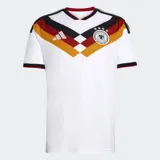 Camiseta de fútbol Adidas de la primera equipación de Alemania, modelo 26. Diseño principal blanco con un patrón en V en el pecho que incorpora los colores de la bandera alemana (negro, rojo y amarillo/dorado). Posee cuello en V con ribete rojo y detalles en las mangas. El escudo de la federación alemana con cuatro estrellas está bordado en el pecho, junto al logo de Adidas.