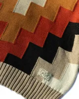 Buzo de tejido de punto con cuello alto y diseño geométrico en zigzag. Presenta una combinación de colores cálidos como naranja, rojo, negro y beige, con puños y cintura acanalados.