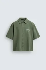 Camisa de corte relajado color verde oliva, con cuello solapa, manga corta y cierre frontal de botones. Presenta un estampado con el texto 'ASTRAL Drift' en el frente.