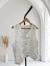 Chaleco blanco con escote en V y cierre frontal de tres botones color hueso. Presenta un tejido texturizado con bordados en relieve de patrón orgánico o floral.