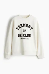 Buzo oversized color crema con cuello redondo acanalado. Presenta un estampado frontal en negro con el texto "VERMONT SKI CLUB Montpelier 1970" y un logo de montaña. Tiene hombros caídos y remates acanalados en puños y bajo.
