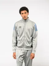 Conjunto deportivo Umbro compuesto por campera de cuello alto con cierre frontal y pantalón de corte recto. La campera presenta bloques de color blanco en los hombros y el logo de la marca bordado en el pecho. Confeccionado en tejido sintético con acabado satinado.