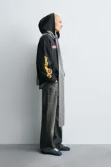 Sudadera tipo hoodie de corte relajado, color negro, con capucha y bolsillo frontal tipo canguro. Presenta parches decorativos en el pecho y estampados en color amarillo en la manga derecha. Acabados en rib en puños y ruedo.