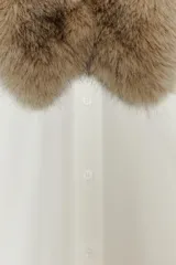 Camisa de popelín color crudo con cuello solapa y aplicación desmontable de pelo sintético en tono marrón. Presenta cierre frontal con botones, manga larga con puños y bajo asimétrico.