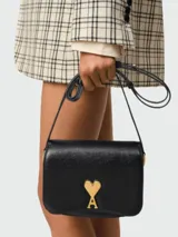 Clutch de cuero negro con solapa frontal y cierre magnético. Presenta el icónico logo metálico dorado en forma de corazón con la letra A en el centro. Incluye una correa fina ajustable y extraíble para llevar al hombro o cruzada.