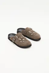 Zueco de cuero descarne color marrón, con diseño estilo western. Presenta una hebilla metálica lateral y múltiples apliques metálicos decorativos y ojales sobre la capellada. Cuenta con suela de goma negra.