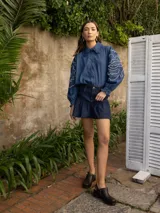 Camisa de denim azul con calce holgado, cuello con volados, mangas largas abullonadas con puños elásticos y bordados decorativos en color crudo. Presenta cierre frontal con botones en forma de flor.