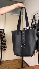Bolso tipo tote bag negro de cuero con textura granulada, con doble asa de hombro y detalle de tira vertical con tachas y aro metálico.