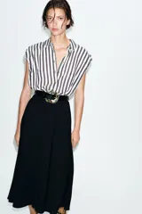 Vestido midi combinado con parte superior de camisa a rayas verticales blancas y negras, cuello solapa, manga sisa con frunces y botones dorados. La parte inferior es una falda midi negra de corte recto. Incluye un cinturón negro con textura de cocodrilo y hebilla dorada.