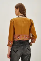 Chaqueta corta color mostaza con mangas abullonadas y bordados étnicos en tonos rosados y rojos en el frente, puños y hombros. Presenta un cierre frontal con detalles de cuentas.