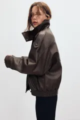 Cazadora tipo biker de cuero sintético en color marrón oscuro, con cuello subido, cierre frontal cruzado con botones forrados y cremallera oculta. Presenta mangas largas con trabillas en los hombros, bolsillos laterales de vivo y bajo con acabado elástico.