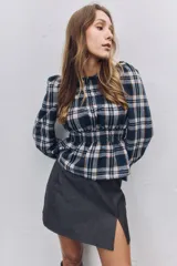 Blusa de manga larga con diseño a cuadros escoceses en tonos azul marino, blanco y naranja. Presenta cuello redondo, hombros ligeramente abullonados y detalle de fruncido elástico en la cintura que crea un efecto peplum.