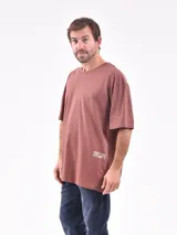 Remera oversize color marrón, de manga corta y cuello redondo, con pequeño estampado tipográfico blanco en el lateral inferior.