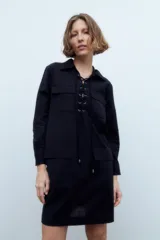 Vestido corto negro con cuello camisero, mangas largas, cierre frontal con cordones y bolsillos de parche en el pecho y las caderas.