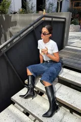 Bota alta de caña media en eco cuero negro, con diseño de punta fina y taco aguja bajo. Presenta un cierre lateral para facilitar el calce y un detalle de costura vertical en el frente.