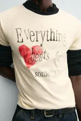 Camiseta de corte regular, color crema, con cuello redondo y ribetes en las mangas y cuello en color negro. Presenta un estampado frontal con un corazón rojo y texto en negro que dice "Everything beats for something".