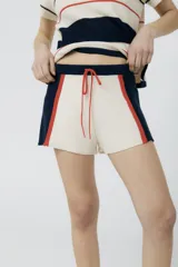 Short tejido en jersey color crudo con cintura elastizada azul marino y cordón de ajuste naranja. Presenta rayas verticales en los laterales con detalles de punto y terminaciones desflecadas.