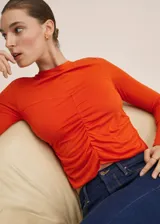 Camiseta de manga larga color naranja, con cuello redondo y diseño entallado. Presenta un detalle fruncido central y costuras decorativas en el frente.