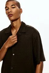 Camisa de manga corta H&M con cuello abierto, confeccionada en tejido de punto con textura. Presenta un diseño de corte holgado con bajo recto y aberturas laterales.