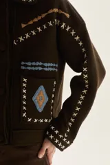 Campera de lana merino hervida en color crudo, con diseño de cuello camisero, cierre frontal de botones y bolsillos frontales. Presenta bordados artesanales con motivos geométricos en tonos marrón y azul en el frente, espalda y mangas.