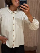 Camisa de algodón color blanco con cuello redondo y detalle de fruncido tipo nido de abeja en el canesú. Presenta cierre frontal con botones decorativos, mangas largas abullonadas con puños elásticos fruncidos y volados en el cuello.