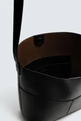 Bolso tipo shopper de cuero sintético negro, con costuras visibles en el frente que dividen el cuerpo en paneles. Presenta un borde superior ondulado y un asa de hombro ajustable.