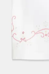 Camiseta de algodón de corte regular, color blanco, con cuello redondo y manga corta. Presenta detalles de bordados florales y ornamentales en color rosado en el ruedo y en las mangas.