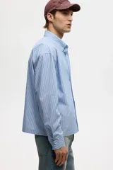 Camisa de corte relajado confeccionada en tejido de algodón con estampado de rayas verticales. Presenta cuello americano, cierre frontal con botones, un bolsillo en el pecho y mangas largas con puños abotonados.