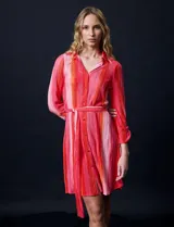 Vestido camisero midi de satén plisado con estampado a rayas verticales en tonos rosa y naranja. Tiene cuello camisero, cierre frontal con botones, mangas largas con puños abotonados y cinturón de lazo.