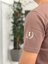 Remera color beige con cuello redondo y mangas cortas. Estampado frontal con el texto "Duxonstudios" en tipografía cursiva, acompañado de un logotipo circular con hojas de laurel y una figura central en tono verde oscuro.