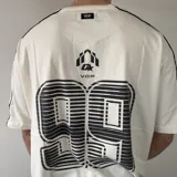 Remera oversize blanca de manga corta con detalles de líneas negras en las costuras de los hombros y mangas. Presenta un gran estampado en la espalda con el número 99 en diseño rayado y el logo de la marca en la parte superior.