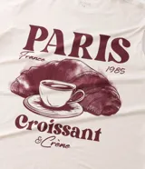 Remera de algodón color crema con cuello redondo y mangas cortas. Presenta un estampado frontal con temática parisina que incluye la palabra 'Paris', un dibujo de una taza de café sobre un croissant y la frase 'Croissant & Crème'.