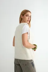 Chaleco de punto con escote en V y cierre frontal de botones. Presenta un diseño de corte recto y textura jaspeada en tono crema.